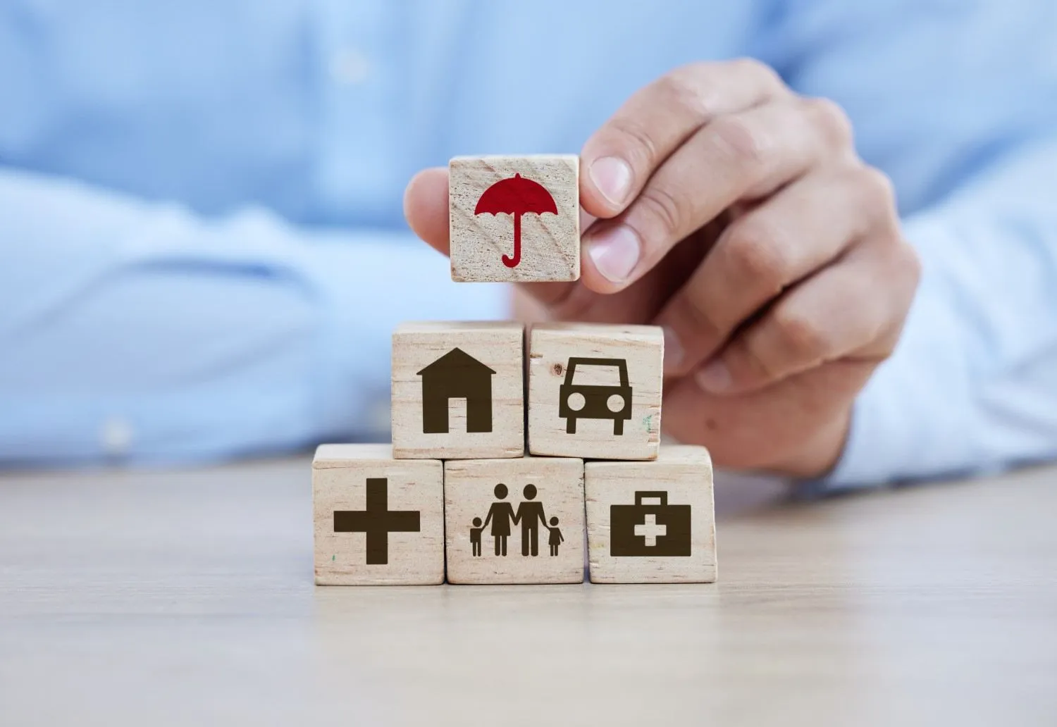 A hand stacking wooden blocks marked with home, car, health and family symbols